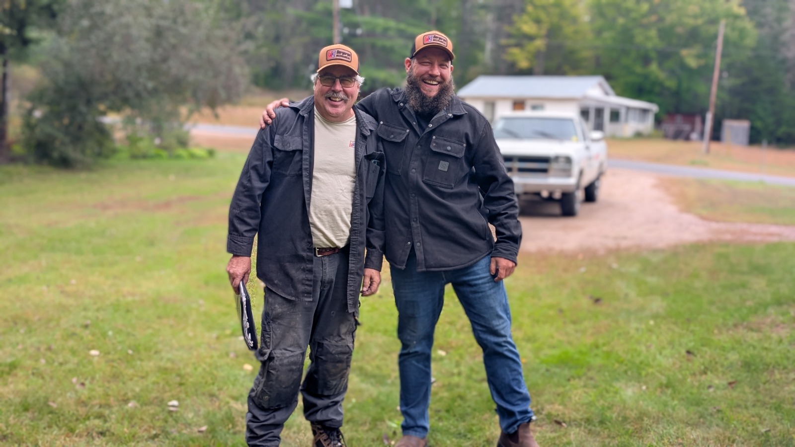 Two members of the Bergeron Mechanical team in company caps, standing on a lawn with a service truck behind them